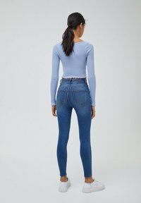 Kvinna med långt mörkt hår i hästsvans, iklädd en ljusblå långärmad crop top, högmidjade blå skinny jeans och vita sneakers, står.