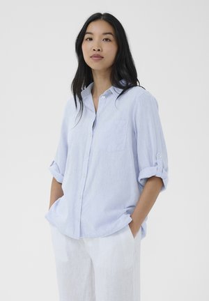 Young woman with long dark hair wearing a light blue button-up shirt with rolled sleeves and white pants, standing with hands in pockets.