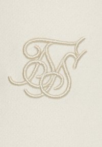 Krämfärgad tyg med en mjuk textur som har en broderad logotyp i ljus beige tråd, med intrikata slingor och virvlar.