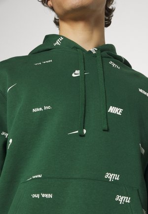 Osoba nosící tmavě zelenou mikinu Nike s bílými logy Nike a textem rozptýleným po celé látce, včetně kapuce a přední kapsy.
