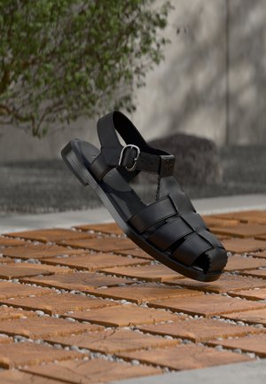 Sandale pour homme en cuir noir avec sangle à boucle flottant au-dessus d'une surface extérieure en pierre et carrelage en bois, avec un fond de feuillage vert et un mur en béton.
