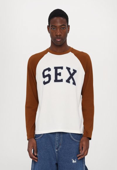Long-sleeve t-shirt with white body and brown raglan sleeves. Features navy text stating "SEX" boldly across the chest. Made of cotton material.