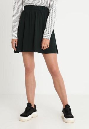 Mørkegrøn skaternederdel med elastik i taljen, kombineret med en hvid langærmet skjorte med striber og sorte sneakers med en chunky sål.