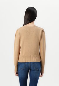 Beige gebreide trui met geribde onderrand en manchetten. Heeft een ronde halslijn en puffy mouwen. Geselecteerd in combinatie met donkerblauwe jeans.