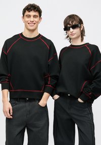 Fekete kapucnis pulóverek piros kontrasztvarrással, raglán ujjakkal. Fekete farmernadrággal párosítva. Két modell áll egymás mellett pózolva.