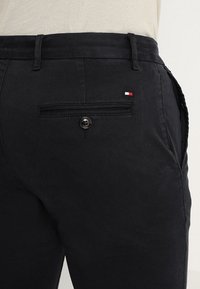 Pantaloni di cotone neri con una tasca posteriore dotata di chiusura a bottone. Piccolo logo ricamato in rosso, bianco e blu vicino alla tasca.