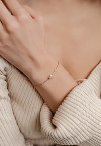 Pulsera en tono dorado con un pequeño colgante en forma de corazón, que presenta una cadena delicada y una textura suave, llevada en una muñeca con un suéter de canalé.