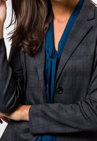 Blazer de cuadros gris en un corte estructurado, con un solo botón negro, combinado con una blusa verde azulado oscuro y texturas de tela suaves.