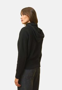 Musta huppari, jossa on teksturoitu neulekuvio, rento leikkaus ja ribattu helma. Yhdistetty tummiin farkkuihin, esittelee rentoa tyyliä.