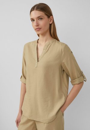 Chemise en lin beige avec un col en V, des manches retroussées et une coupe décontractée. La texture semble douce avec des détails de couture minimaux.