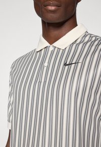 Svītrainā polo krekls krēmkrāsā un tumši pelēkā, ar pogājamu apkakli un mazu melnu Nike logo kreisajā krūtī. Gluda auduma tekstūra.