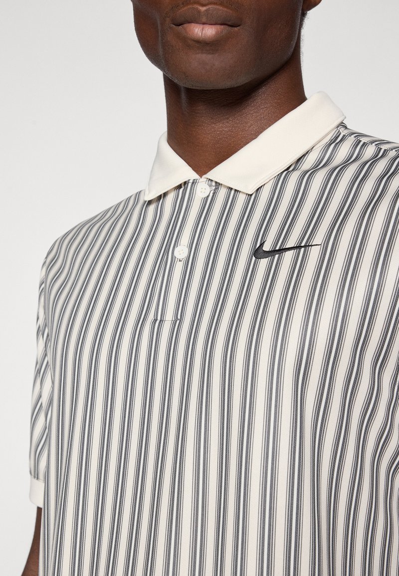 Polo a righe in crema e grigio scuro, con colletto a bottoni e un piccolo logo Nike nero sul lato sinistro del petto. Tessuto dalla texture liscia.