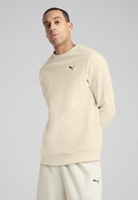 Sudadera beige de cuello redondo con mangas raglán, logo negro de Puma en el pecho, textura de tela suave, combinada con pantalones de color claro.