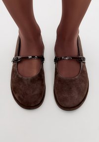 Scarpe Mary Jane in suede marrone con una cintura nera lucida che attraversa il metatarso, punta arrotondata e una superficie morbida e testurizzata.