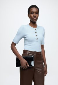 Ανοιχτό γαλάζιο ριμπ πλεκτό τοπ με κοντά μανίκια και πέντε χρυσά κουμπιά· κρατώντας μαύρο τσαντάκι από δερματίνη λουστρίνι· καφέ παντελόνι από συνθετικό δέρμα.