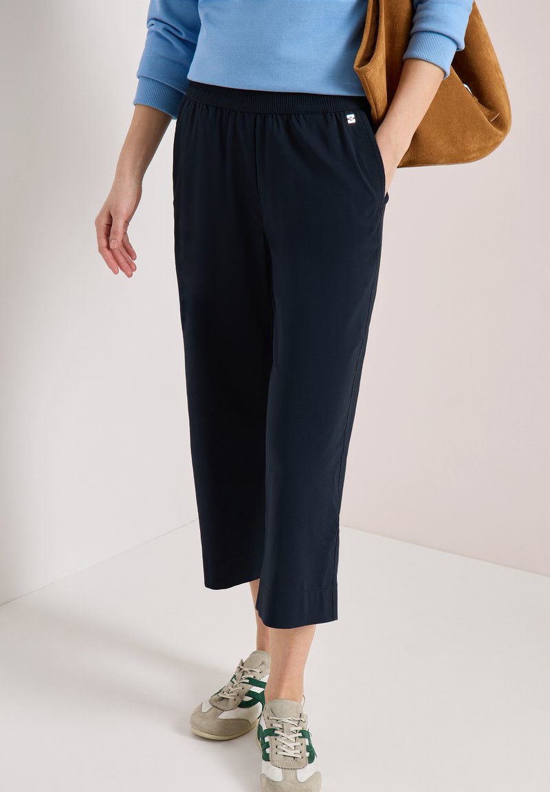 Osoba w granatowych spodniach 7/8, niebieskiej bluzce z długim rękawem, beżowych sneakersach z zielonymi akcentami i z brązową zamszową torbą na ramieniu.