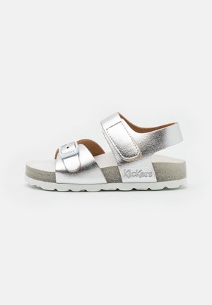 Sandalias metálicas plateadas con una tira ancha, hebilla ajustable y plantilla texturizada. Presenta una suela blanca con un sutil acento gris.