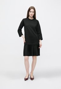 Młoda kobieta stojąca, ubrana w czarną bluzkę z długim rękawem i plisowaną czarną spódnicę, w parze z ciemnobrązowymi spiczastymi butami, na jednolitym jasnym tle.