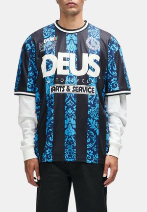 Hombre que lleva una camiseta de manga corta a rayas negras y azules con mangas largas blancas debajo, con un gran texto blanco "DEUS" y patrones decorativos.