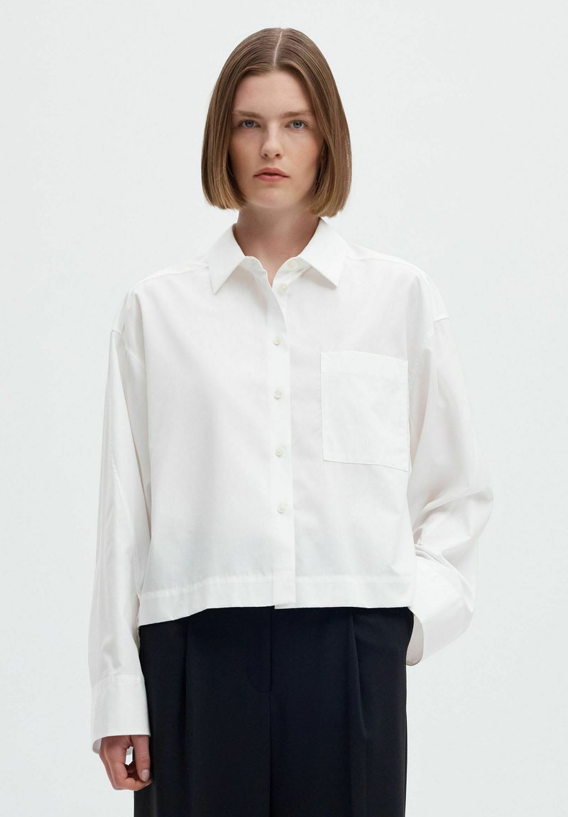 Jeune personne aux cheveux lisses mi-longs, portant une chemise blanche ample boutonnée et un pantalon noir sur un fond uni.