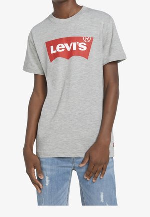 Person iført en lysegrå Levi's t-shirt med et rødt logo og lyseblå revnede jeans, stående med hænderne nær lårene.