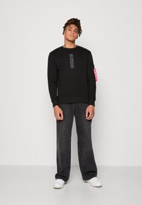 Sudadera negra con una franja de logo en el centro, combinada con pantalones anchos de mezclilla gris oscuro y zapatillas blancas. Presenta una etiqueta lateral.