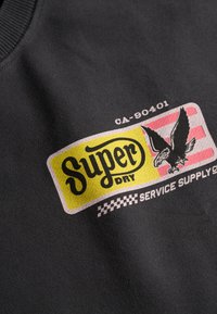 Sudadera gris oscuro con un logo colorido que incluye "Super Dry", un diseño de águila y patrones a cuadros. Tejido suave con cuello acanalado.