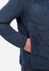 Ręka w bocznej kieszeni granatowej polarowej kurtki o teksturowanej, puszystej tkaninie i gładkiej listwie zamka błyskawicznego po bokach.