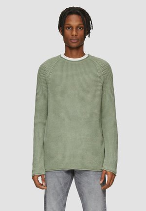 Groene gebreide trui met een gestructureerd patroon, ronde halslijn en lange mouwen. Draagt over een witte blouse, gecombineerd met lichtgrijze jeans.