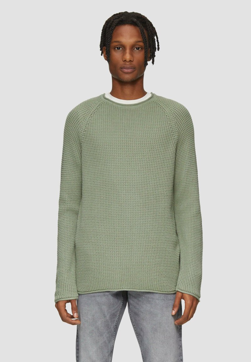 Grüner Strickpullover mit strukturiertem Muster, Rundhalsausschnitt und langen Ärmeln. Über einem weißen Hemd getragen, kombiniert mit hellgrauen Jeans.
