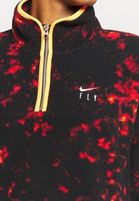 Personne porte un pull polaire noir avec un motif rouge vif, une doublure de fermeture éclair orange et un swoosh Nike blanc avec le texte "FLY" sur la poitrine.