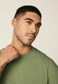 Vihreä puuvillainen t-paita, jossa pyöreä pääntie. Malli pitää kangasta kauluksen läheltä esitellen sileää rakennetta ja vartalonmyötäistä mallia.