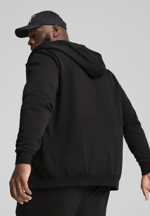Sort hættejakke lavet af blødt materiale med hætte, subtil tekstur og en afslappet pasform. Fremhævet med en sort kasket for et koordineret look.