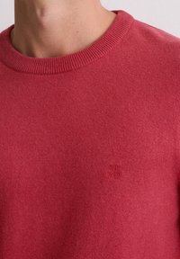 Czerwony sweter z dzianiny z okrągłym dekoltem i subtelną fakturą. Na piersi małe haftowane logo. Miękki materiał o średniej grubości.