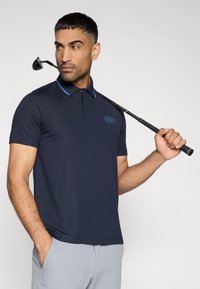 Μπλε σκούρο πουκάμισο πόλο με κοντά μανίκια, με γαλάζιο γιακά και λογότυπο EA7, συνδυασμένο με ανοιχτό γκρι παντελόνι, χειρισμένο τον γιακά από έναν άνδρα με μια ράβδο γκολφ.