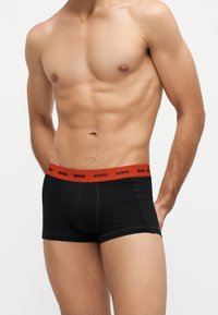 Homme torse nu portant un boxer noir avec une ceinture rouge portant le logo Diesel, debout devant un fond blanc uni.