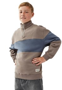 Tonårspojke med kort röd hår som bär en brun och bl�å sweatshirt med kvart dragkedja och grå jeans, står med händerna på höfterna och ler.