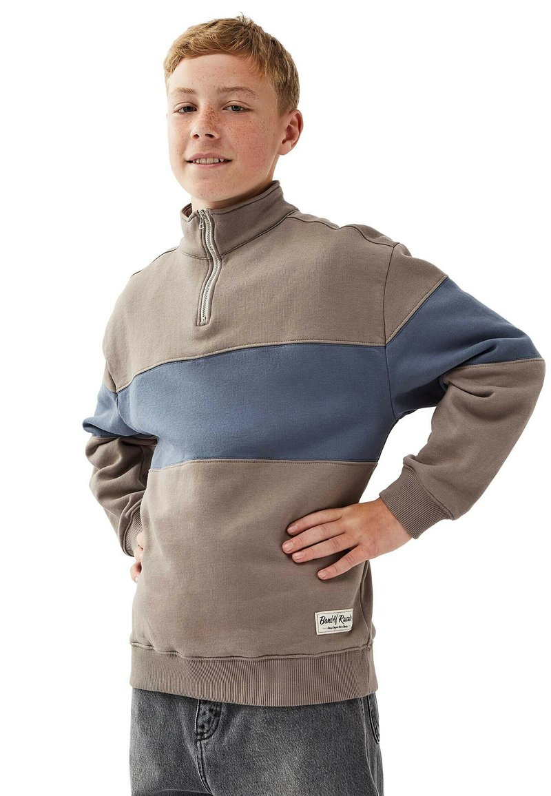 Tonårspojke med kort röd hår som bär en brun och blå sweatshirt med kvart dragkedja och grå jeans, står med händerna på höfterna och ler.