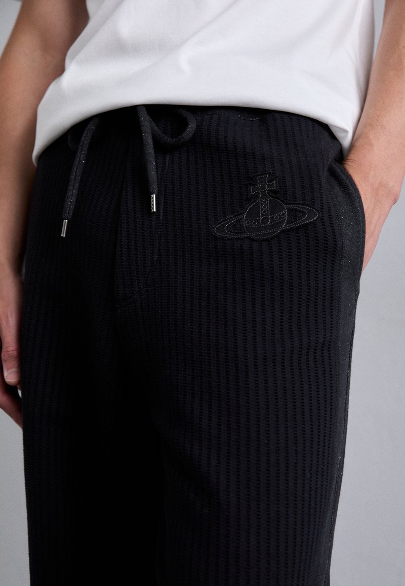 Pantalon de survêtement noir à texture gaufrée avec une taille élastique et un cordon de serrage. Présente un accent graphique brodé subtil sur la cuisse.
