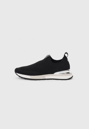Sort slip-on strikket sneaker med hvid sål og sølv accent på hælen, vist på neutral grå baggrund vendt mod venstre.