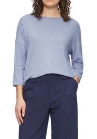 Vrouw gekleed in een lichtblauwe trui met lange mouwen, in de taille van een hoge taille marineblauwe broek gestoken, met een hand in een zak.