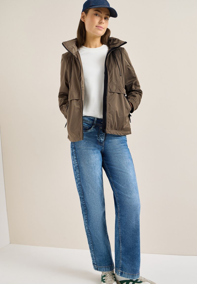 Femme portant une veste marron sur une chemise blanche, un jean bleu, des baskets vertes et blanches, et une casquette bleu marine, debout les mains dans les poches.