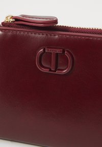 Pochette in pelle bordeaux con finitura testurizzata, dotata di una cerniera dorata e di un logo in rilievo ben visibile sul davanti.