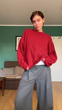 Roter, übergroßer Pullover mit gerippten Bündchen und Saum, kombiniert mit grauen, weit geschnittenen Hosen mit Falten. Glatte Stofftexturen erkennbar.