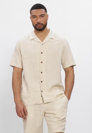 Mand med kort hår og skæg iført en beige kortærmet skjorte med knapper og matchende shorts, stående med den ene hånd i lommen.