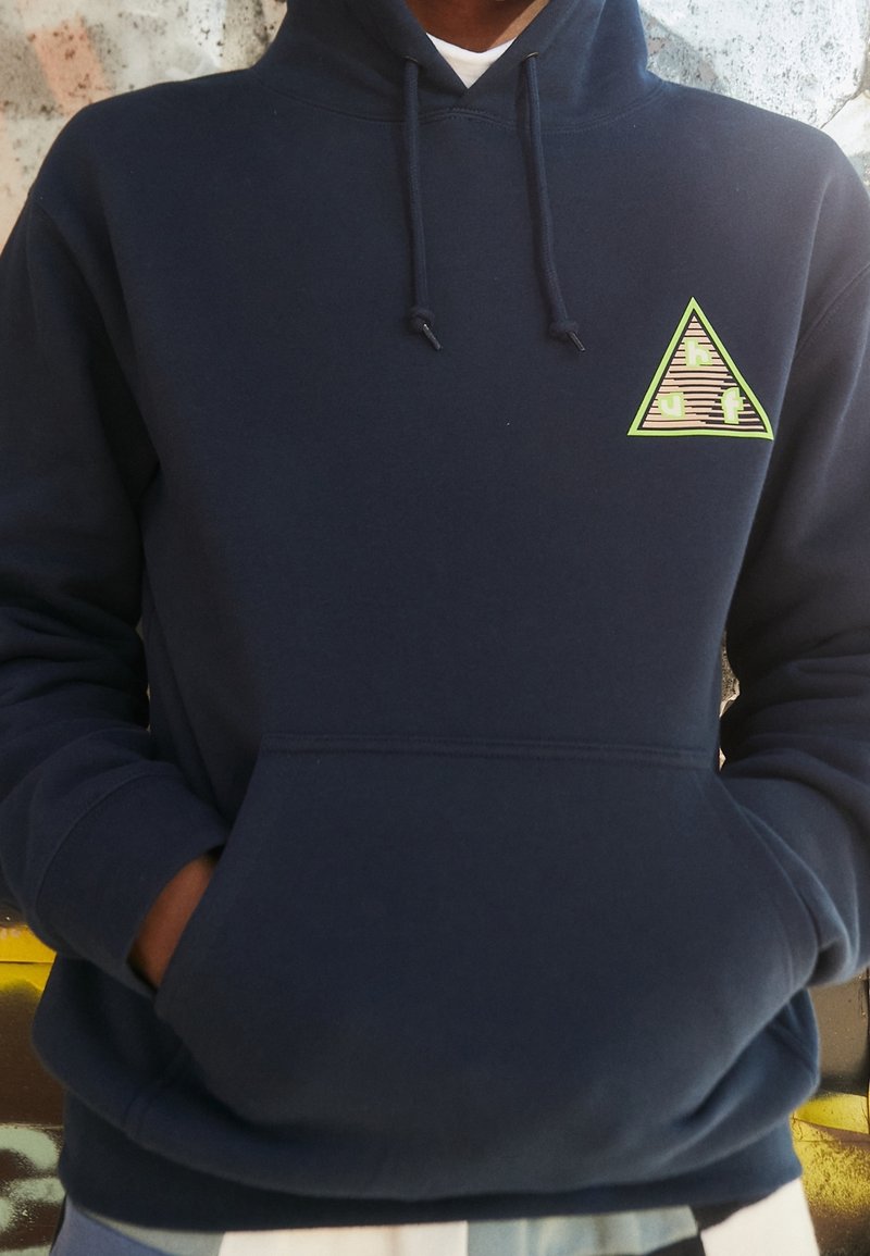 Sudadera con capucha azul marino con bolsillo canguro y un logotipo triangular de acento en verde y tela estampada en el área del pecho izquierdo.