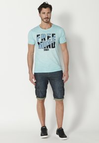 Világoskék, rövid ujjú póló fekete "FREE MIND 2001" felirattal, sötét farmer cargo rövidnadrággal és fekete sportcipővel párosítva.