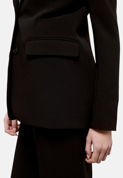 Blazer nero su misura con un design strutturato, chiusura a un bottone, texture liscia e un dettaglio di tasca frontale nascosta.