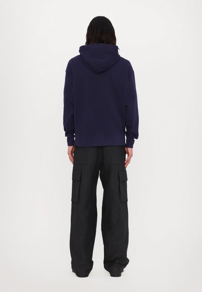 Felpa con cappuccio di colore navy con polsini a costine, abbinata a pantaloni cargo neri con tasche laterali e vestibilità ampia. Modello di spalle.
