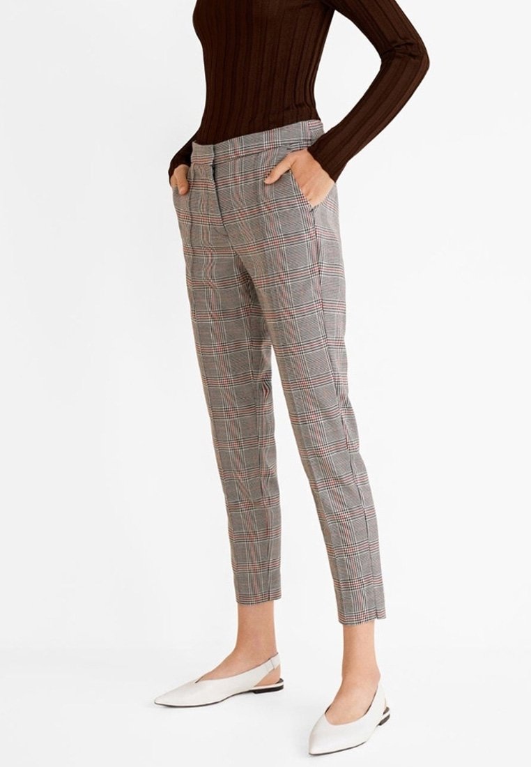 Femme portant un pantalon slim court à carreaux gris, un haut marron en maille côtelée à manches longues et des ballerines blanches à bride arrière, debout les mains dans les poches.